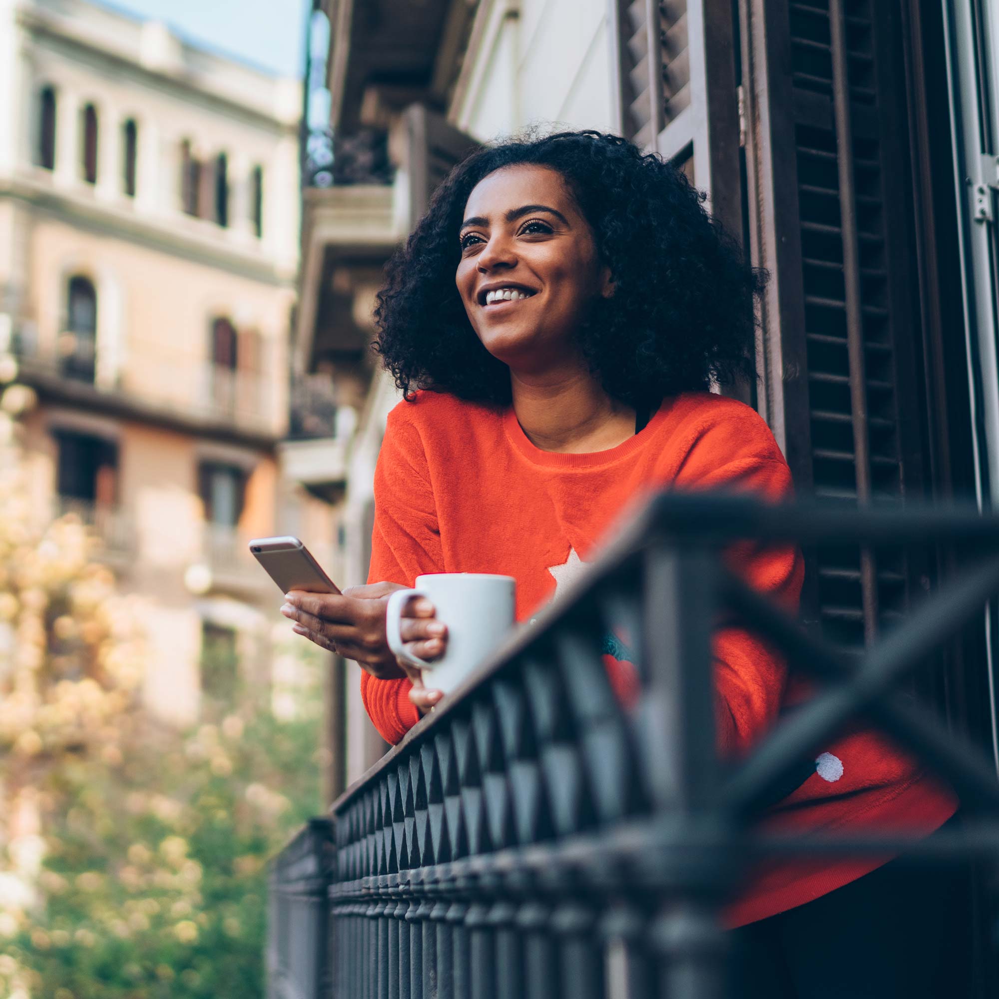 A woman in an orange shirt looks out from the balcony with a cup of coffee in hand.